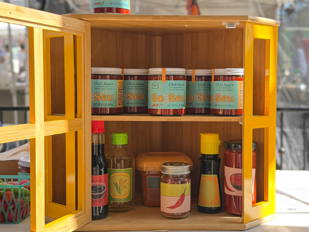 A wooden spice cabinet with yellow-painted side panels, filled with jars of chili sauces and condiments on a tabletop.